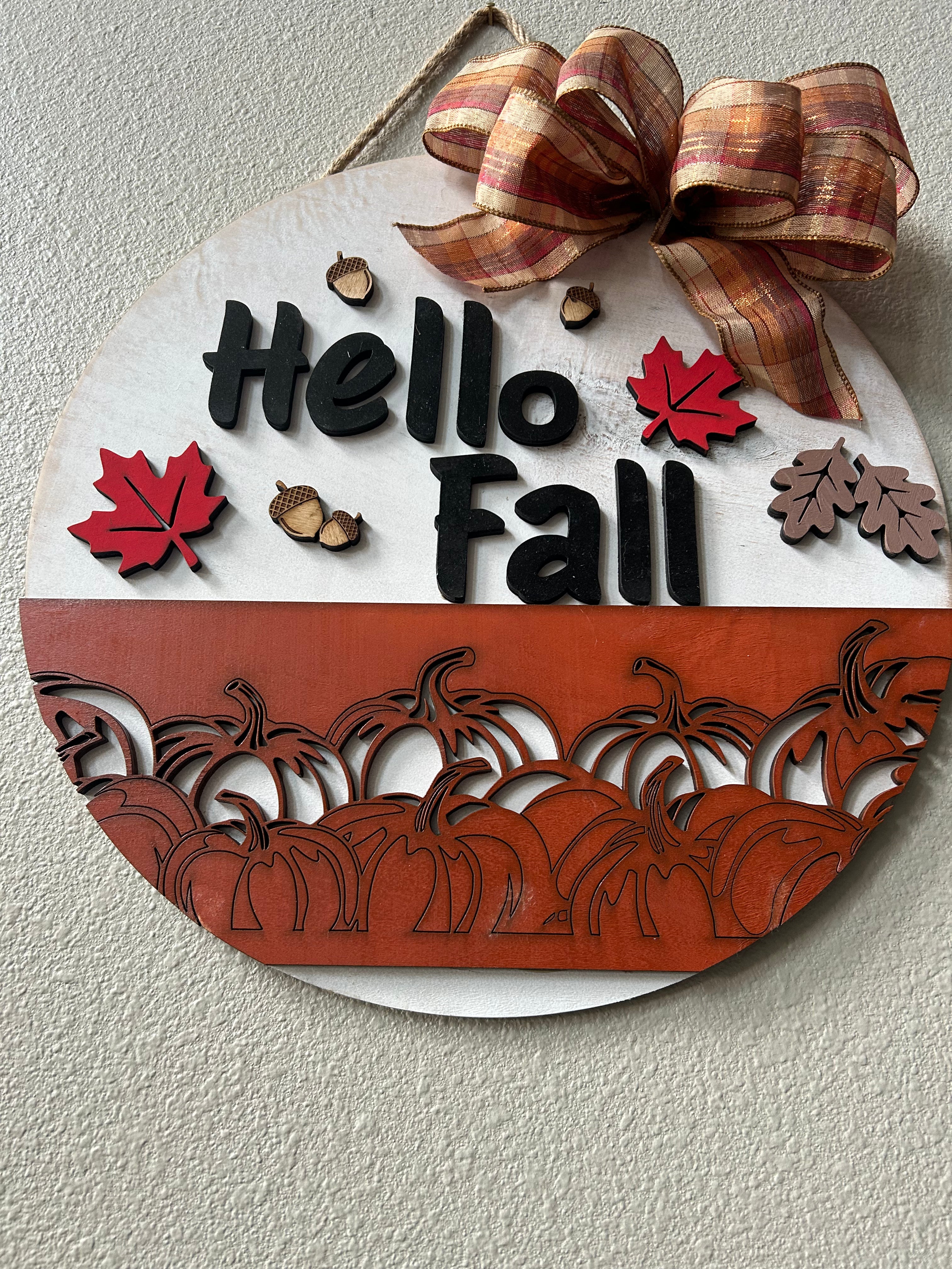 Round fall-themed wall hanging with a white top half reading “Hello Fall” in black letters, a red-orange bottom half with laser-cut pumpkins, a plaid brown ribbon bow, and small red and brown autumn accents, shown mounted on a textured beige wall.